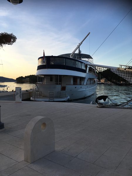 A white superyacht moored stern-first at a stone quay at sunset, with calm water and a wooded headland behind