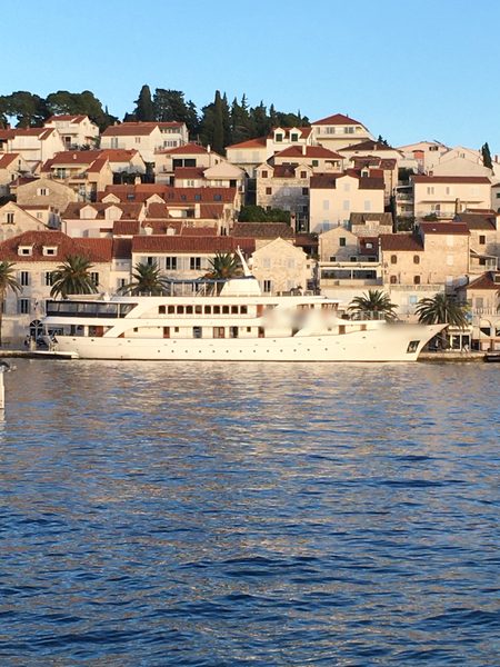 A long white superyacht moored in front of a hillside town of stone houses with red roofs and palm trees